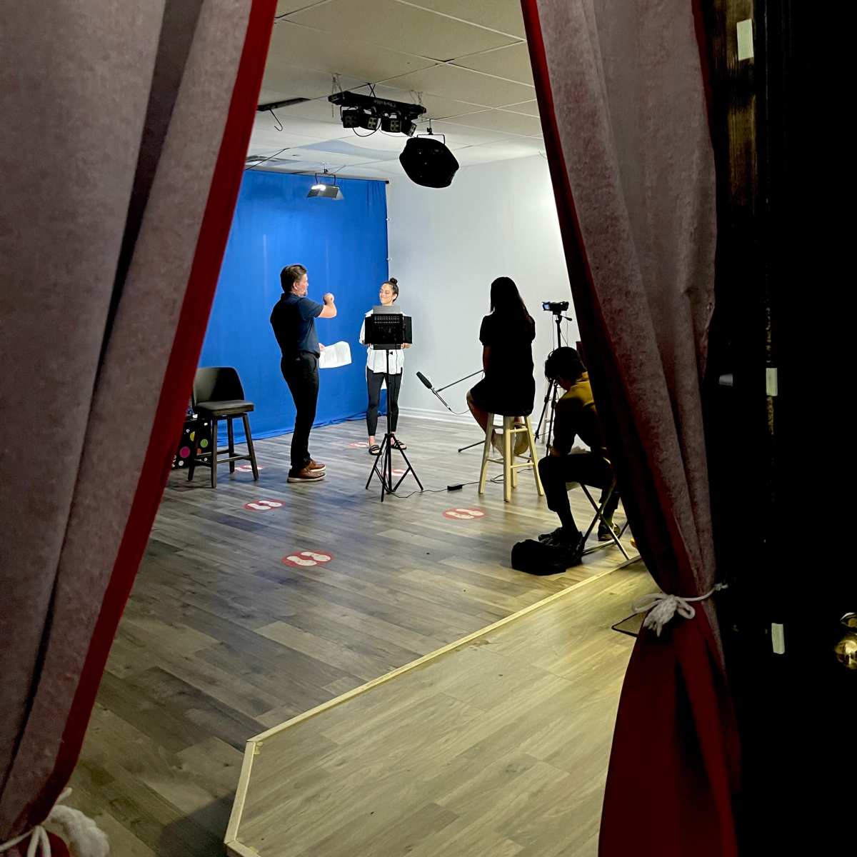 A small stage is framed by red curtains, with two people standing in front of a blue backdrop while others sit and observe.