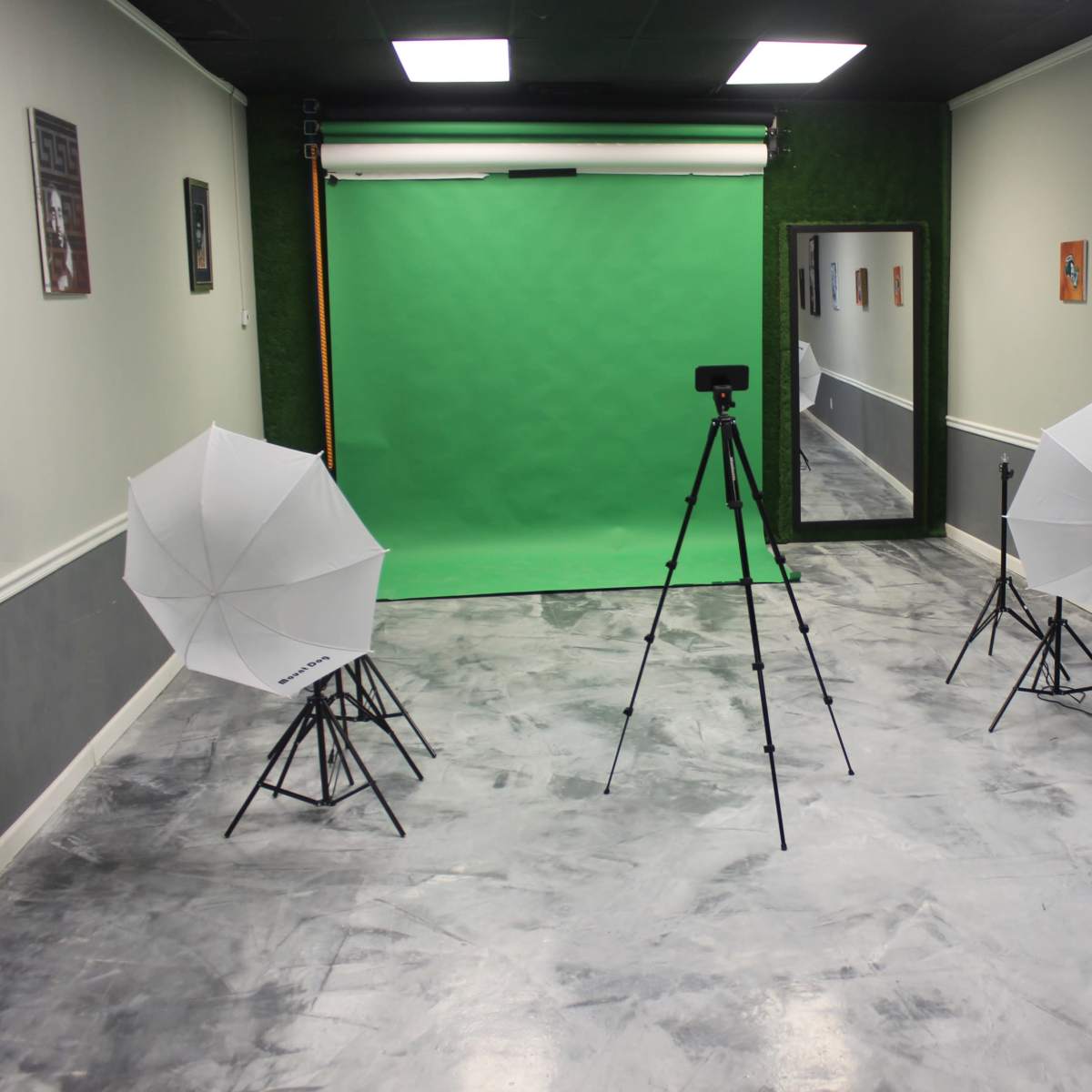 A photography studio setup with a green backdrop, two softbox lights on tripods, and a camera on a tripod in a sparsely decorated room.