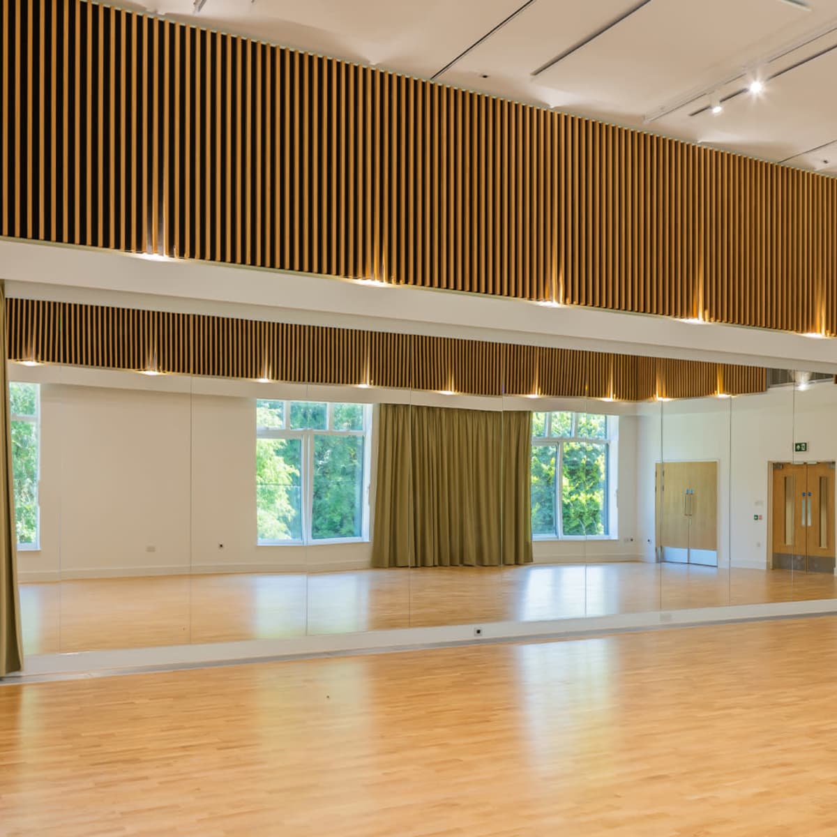 A spacious room with wooden paneling, large mirrors, and natural light coming through windows, featuring a wooden floor and green curtains.