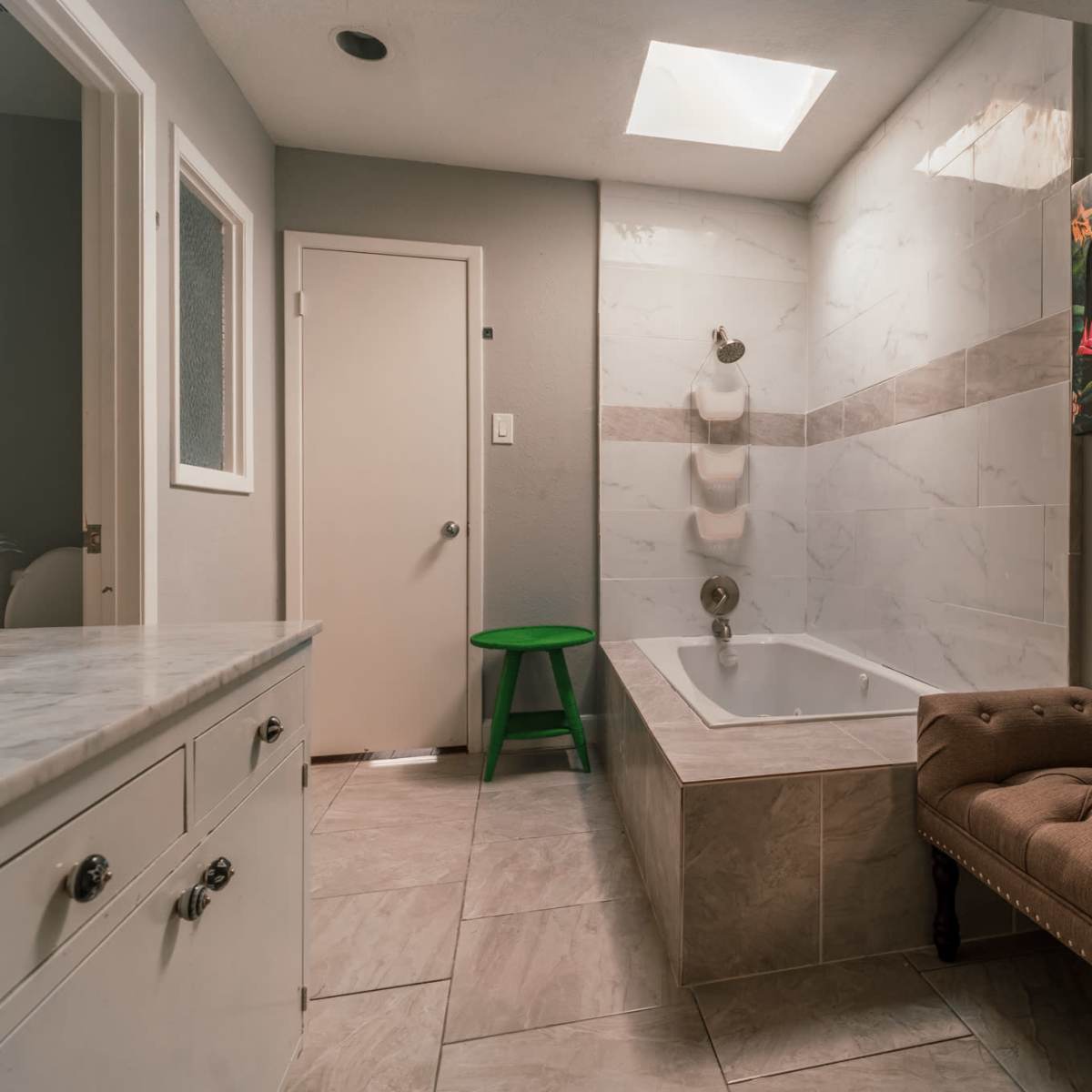 A bathroom with a bathtub, a patterned artwork on the wall, a green stool, and a vanity with drawers.