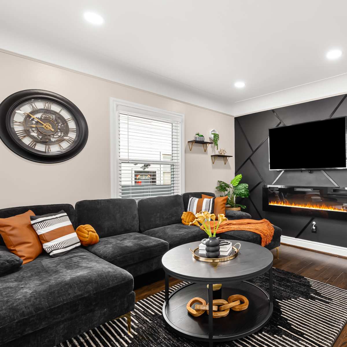 The living room features a black sectional sofa, a round coffee table with a decorative chain, and an accent wall with a mounted TV and a modern electric fireplace.