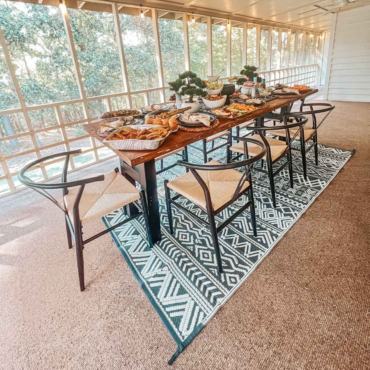 A wooden dining table set with a variety of food surrounded by chairs, located in a sunlit room with large windows.