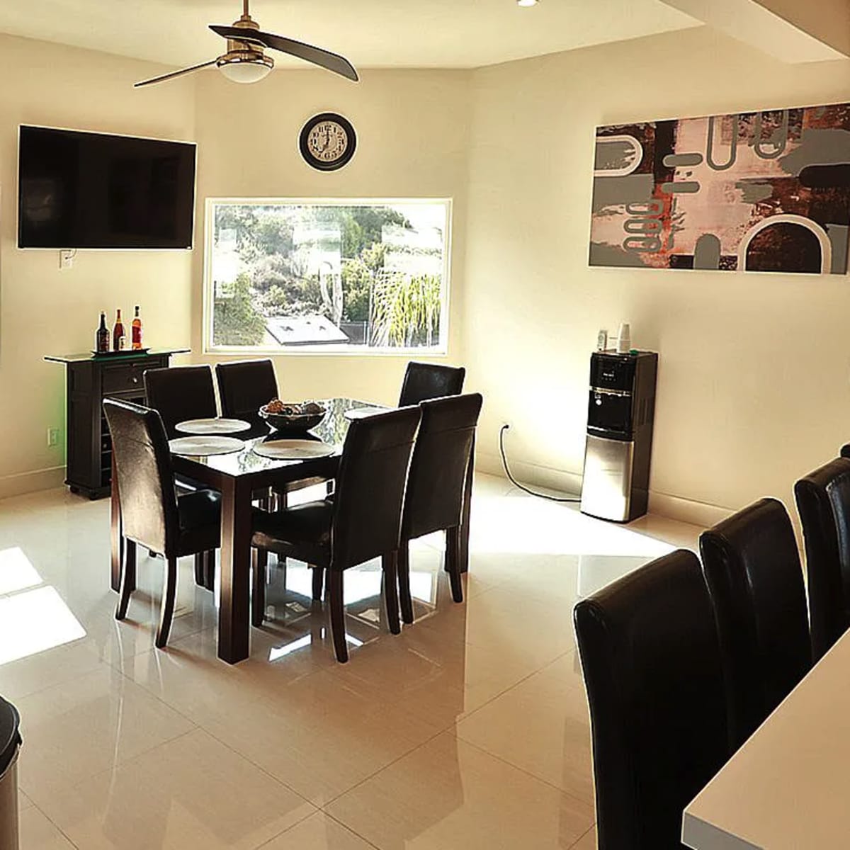 A dining area features a table with eight chairs, a television mounted on the wall, and a window overlooking a garden.