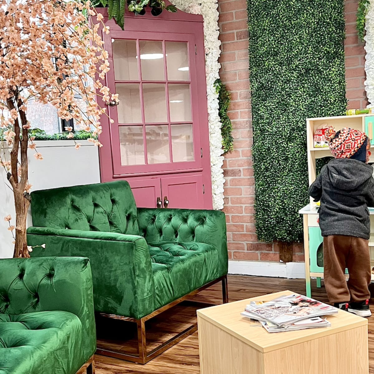 A cozy interior featuring two green velvet chairs, a wooden coffee table, and a child playing at a small activity board, with decorative plants and a pink door in the background.