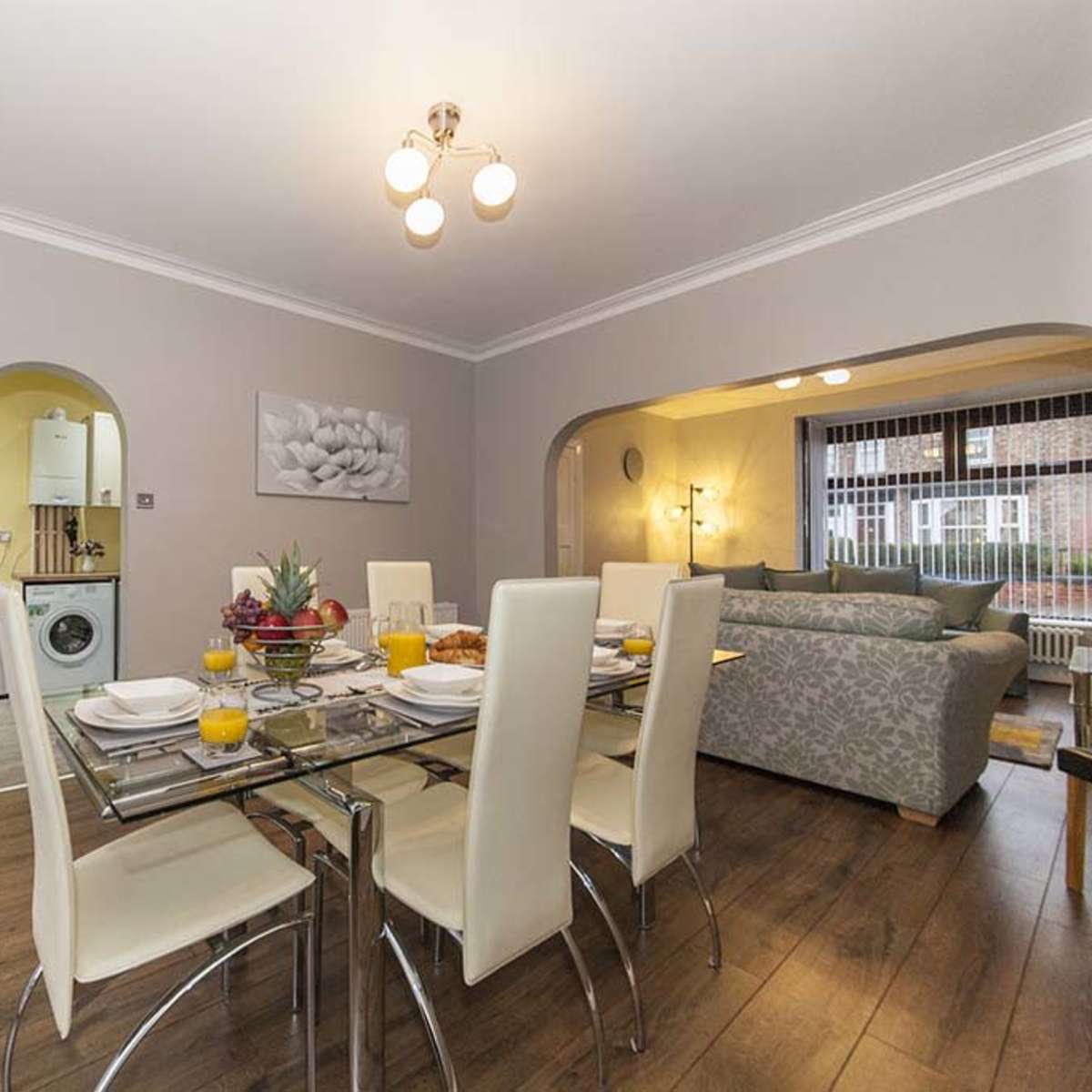 A dining area features a glass table set for a meal, with chairs around it and an open view to a living space beyond.