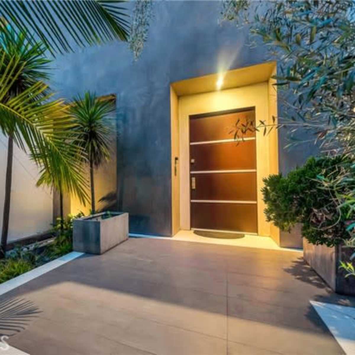 A modern entrance with a wooden door framed by tropical plants and a well-lit pathway.