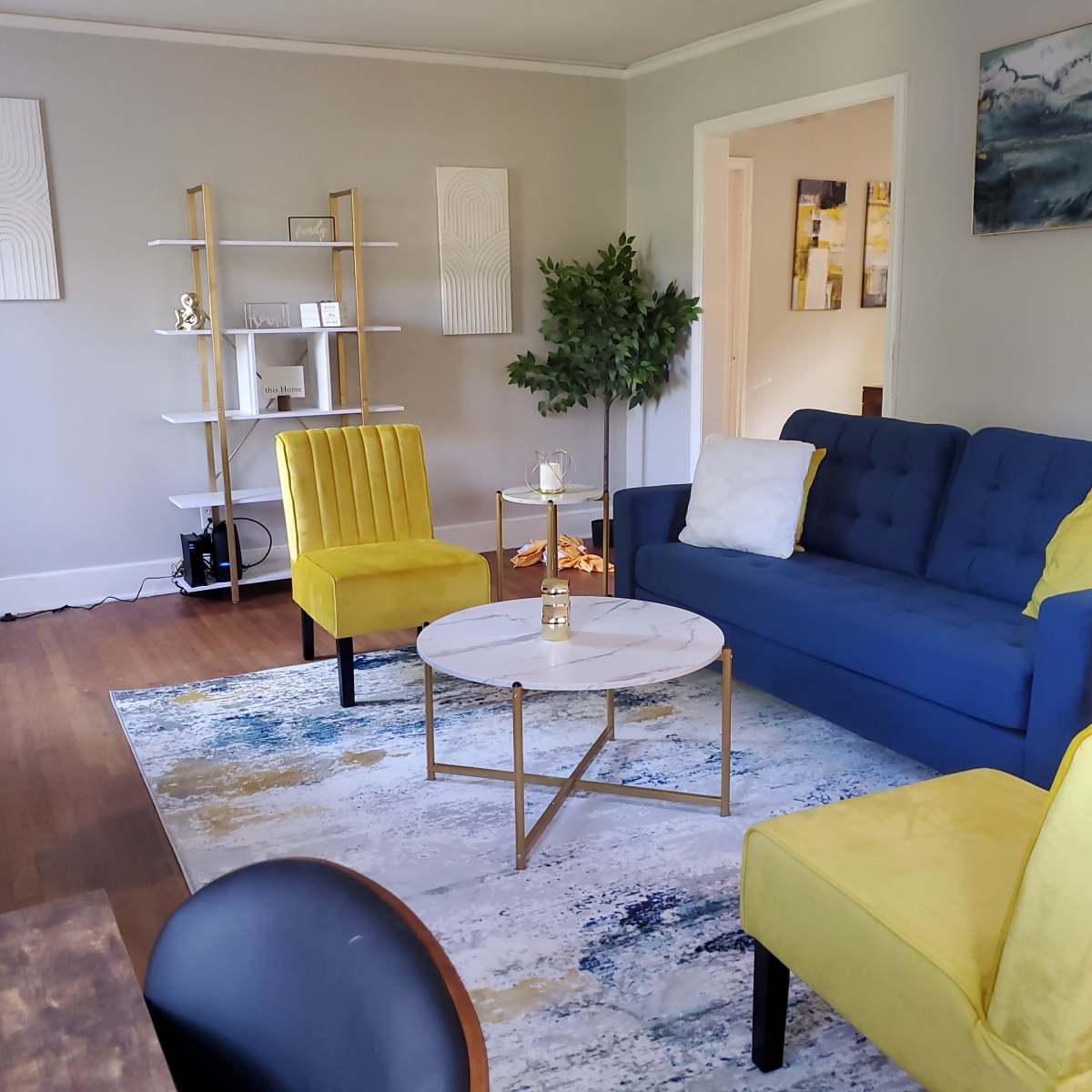 The living room features a blue sofa, two yellow chairs, a round coffee table, and a decorative rug, all arranged around a bookshelf and a potted plant.