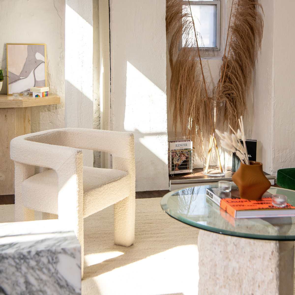 A bright, minimalist living space with a textured chair, a marble coffee table, and decorative elements like pampas grass and books.