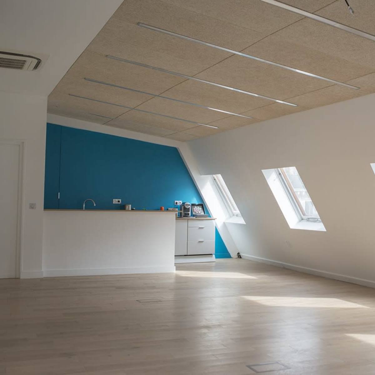 A spacious, well-lit room with angled windows and a blue accent wall, featuring a small kitchenette along one side.
