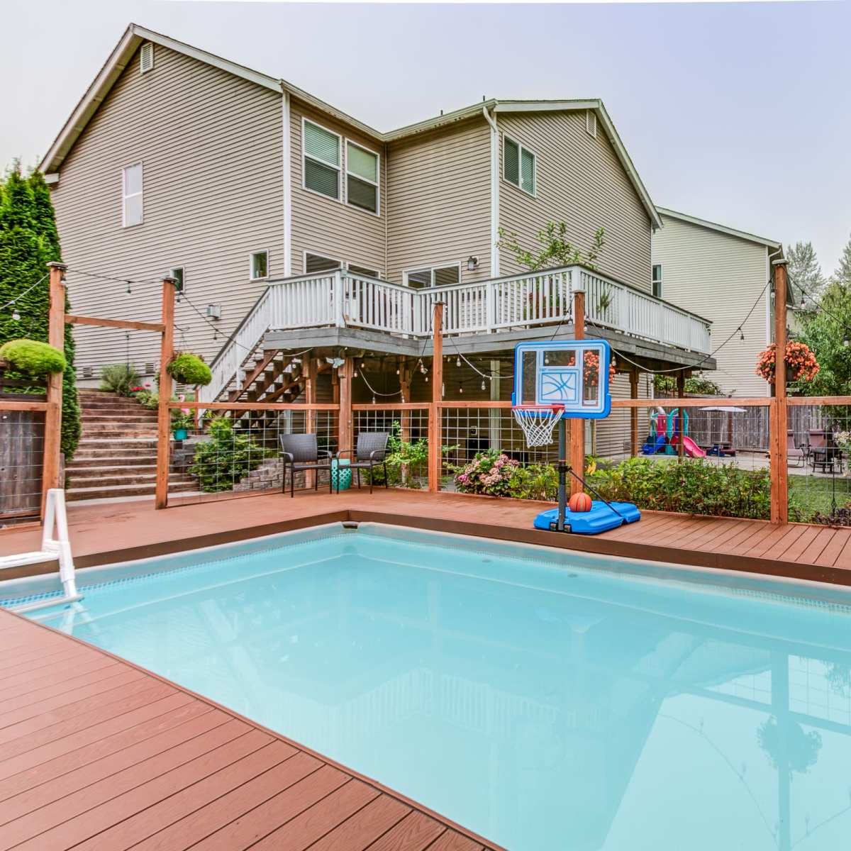 A backyard features a swimming pool surrounded by a wooden deck and landscaped area, with a house and children's play equipment visible in the background.