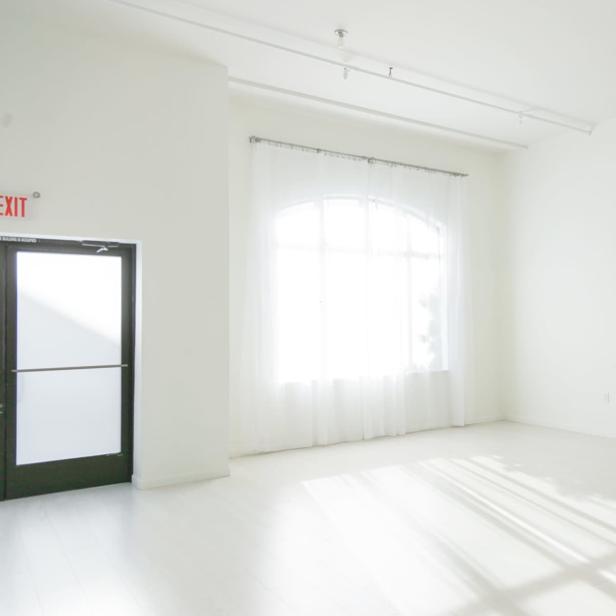 A minimally furnished, bright room with large windows, white walls, and light-colored flooring.