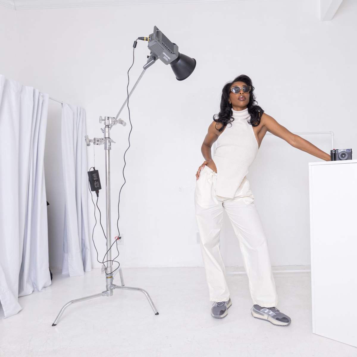 A person in a white outfit poses confidently next to a white pedestal and photography equipment in a bright studio space.