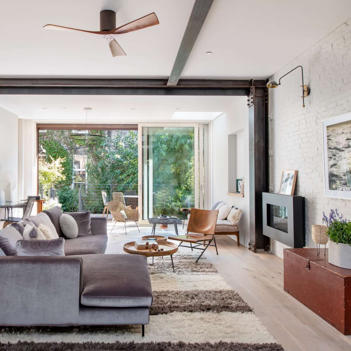 A modern living room features a gray sectional sofa, a wooden coffee table, and large windows that open to a balcony with greenery outside.