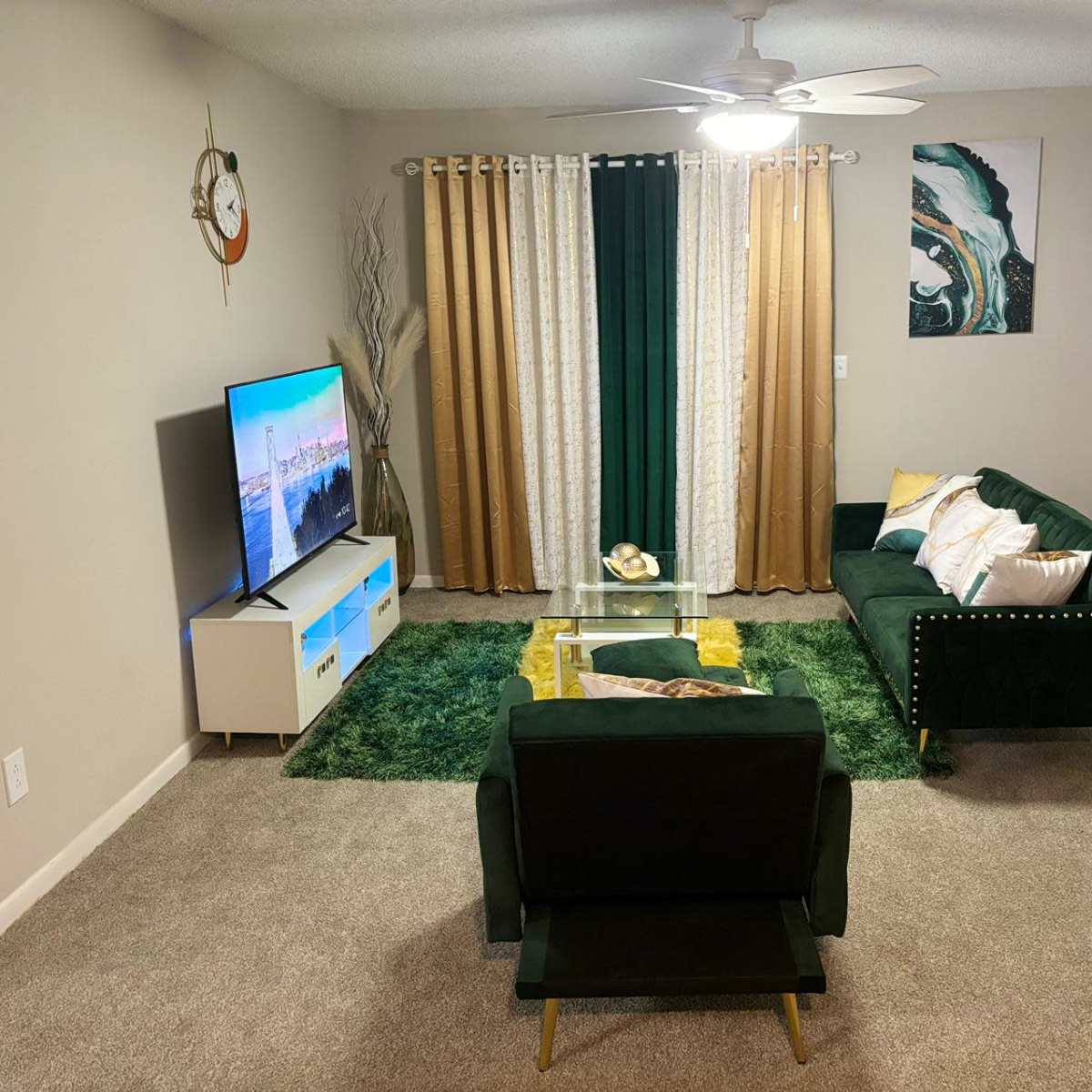 The living room features a green couch, a matching armchair, a glass coffee table, and a large television against a wall decorated with curtains and wall art.