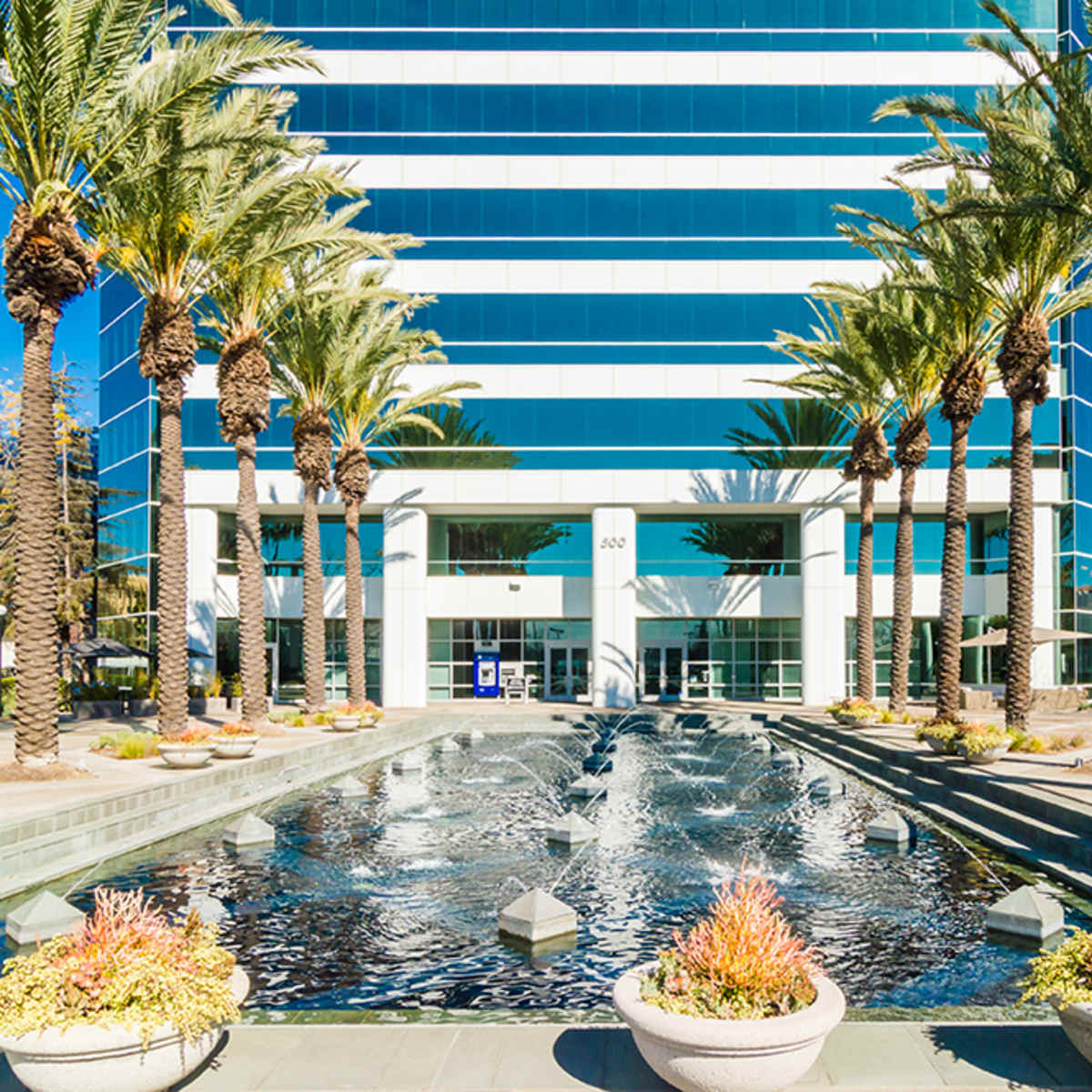 A modern office building is flanked by palm trees and features a decorative water fountain in the foreground.