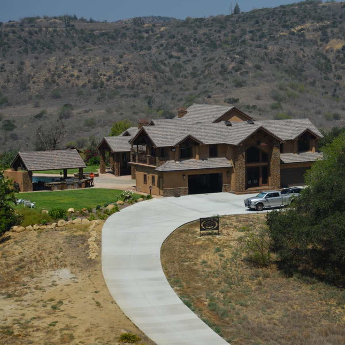 A large, multi-story house with a circular driveway, surrounded by sparse landscaping and rolling hills in the background.