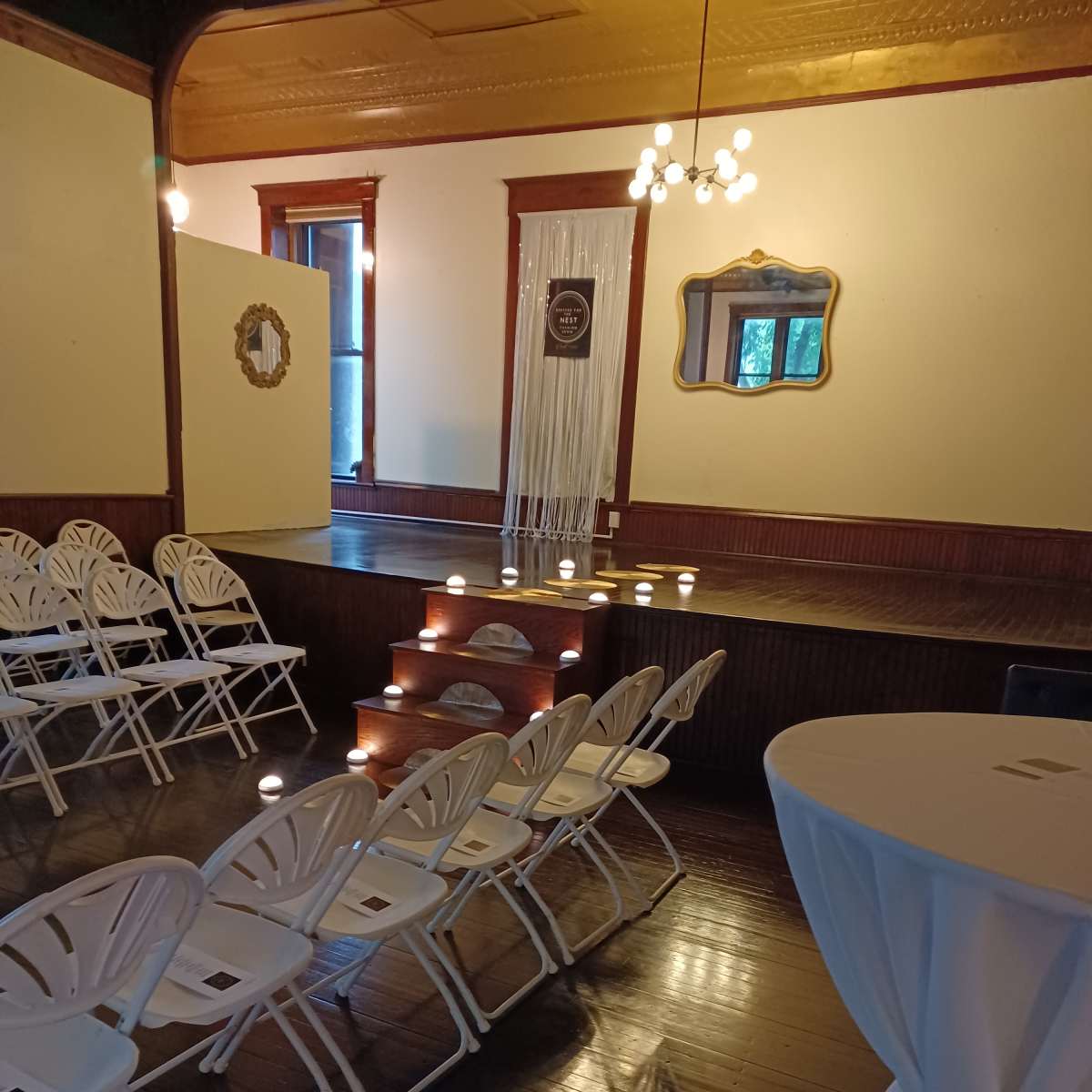 A softly lit event space with white folding chairs arranged facing a small stage, which features a backdrop with string lights and candles along the steps.