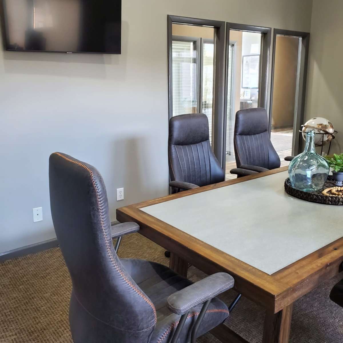 A conference room features a wooden table surrounded by three chairs, with a television mounted on the wall and mirrors reflecting the space.
