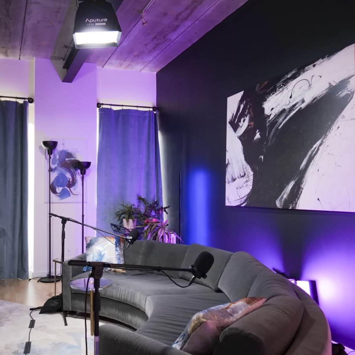 A filming setup is arranged in a living space featuring a couch, lighting equipment, and a large abstract painting on the wall.