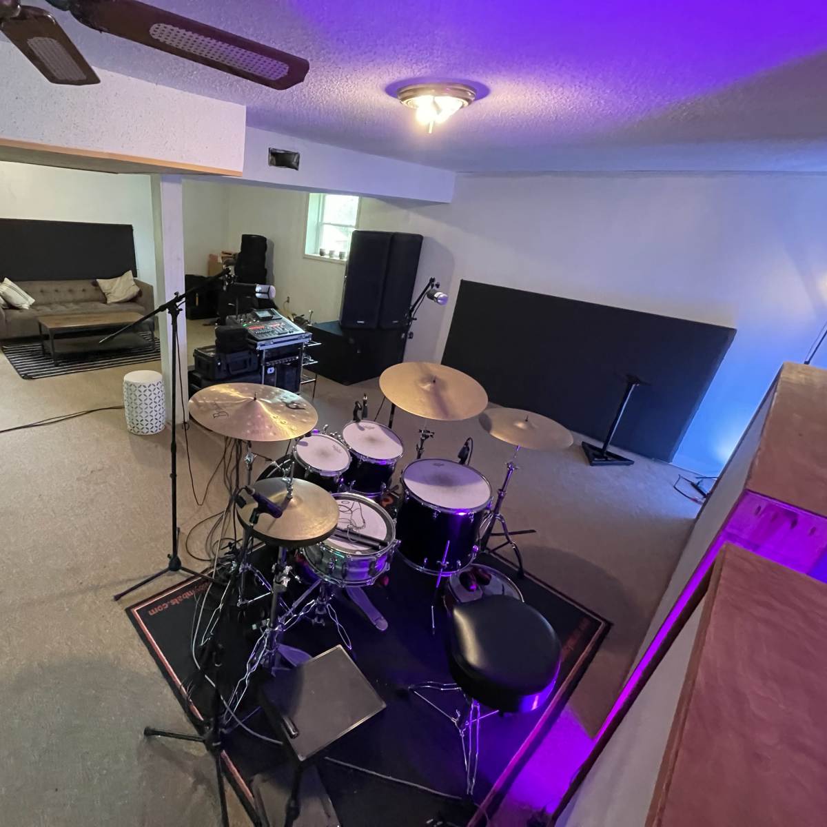 A spacious room featuring a drum set, a sofa, and sound equipment, with a purple-lit ambiance.