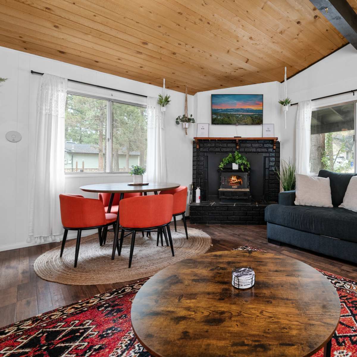 A cozy living room with a dark sofa, a round wooden coffee table, and a dining area featuring a round table with red chairs, all set against a backdrop of large windows and a fireplace.