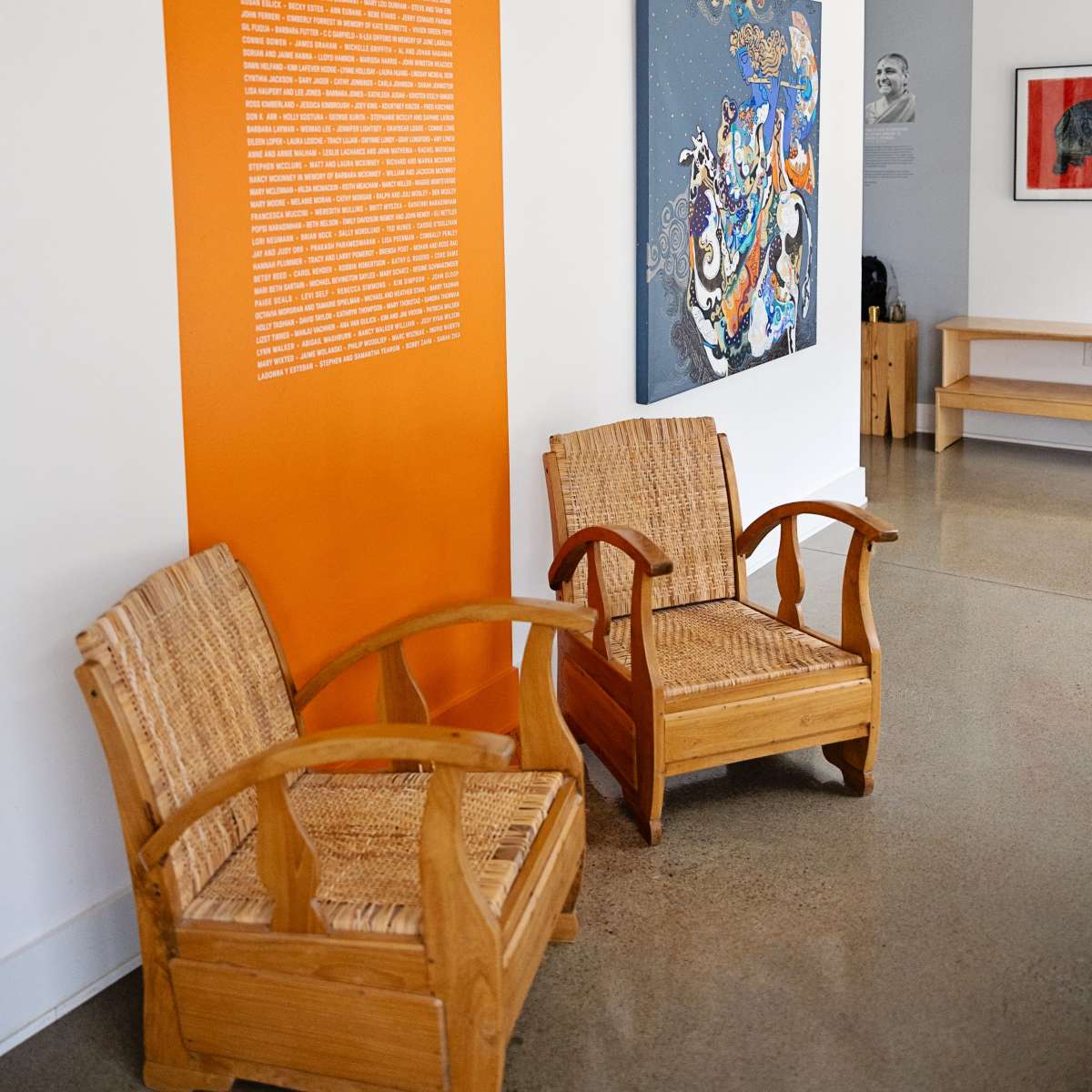 A modern interior space with two wooden chairs, an orange wall featuring text, and colorful artwork hanging nearby.