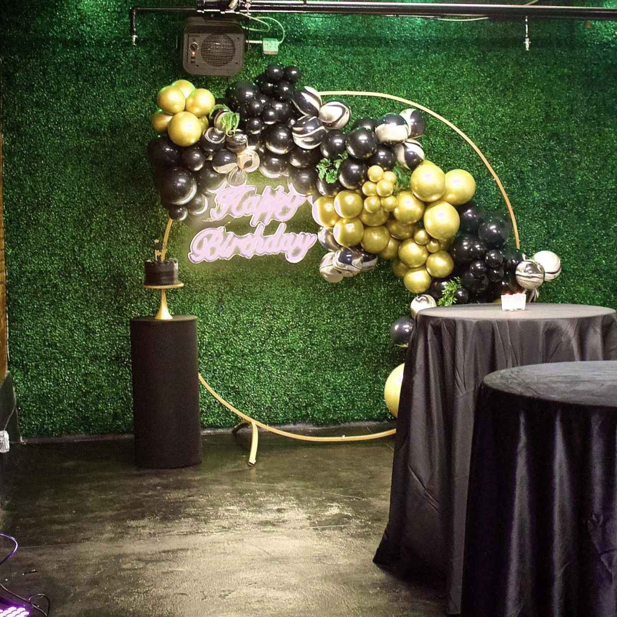 A decorated event space featuring a green grass wall adorned with black and gold balloons, a neon "Happy Birthday" sign, and two black tablecloth-covered tables.