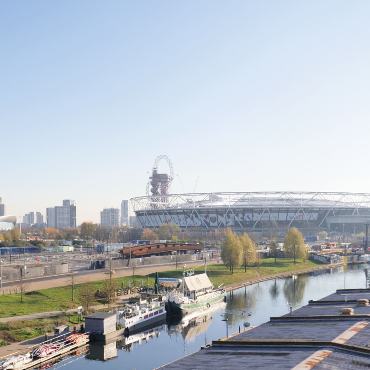 A panoramic view of a stadium surrounded by modern buildings, with a canal and boats in the foreground.