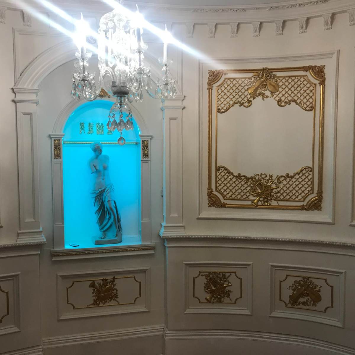 An ornate interior featuring a chandelier and a niche with a statue illuminated by blue light, framed by decorative gold accents on the walls.