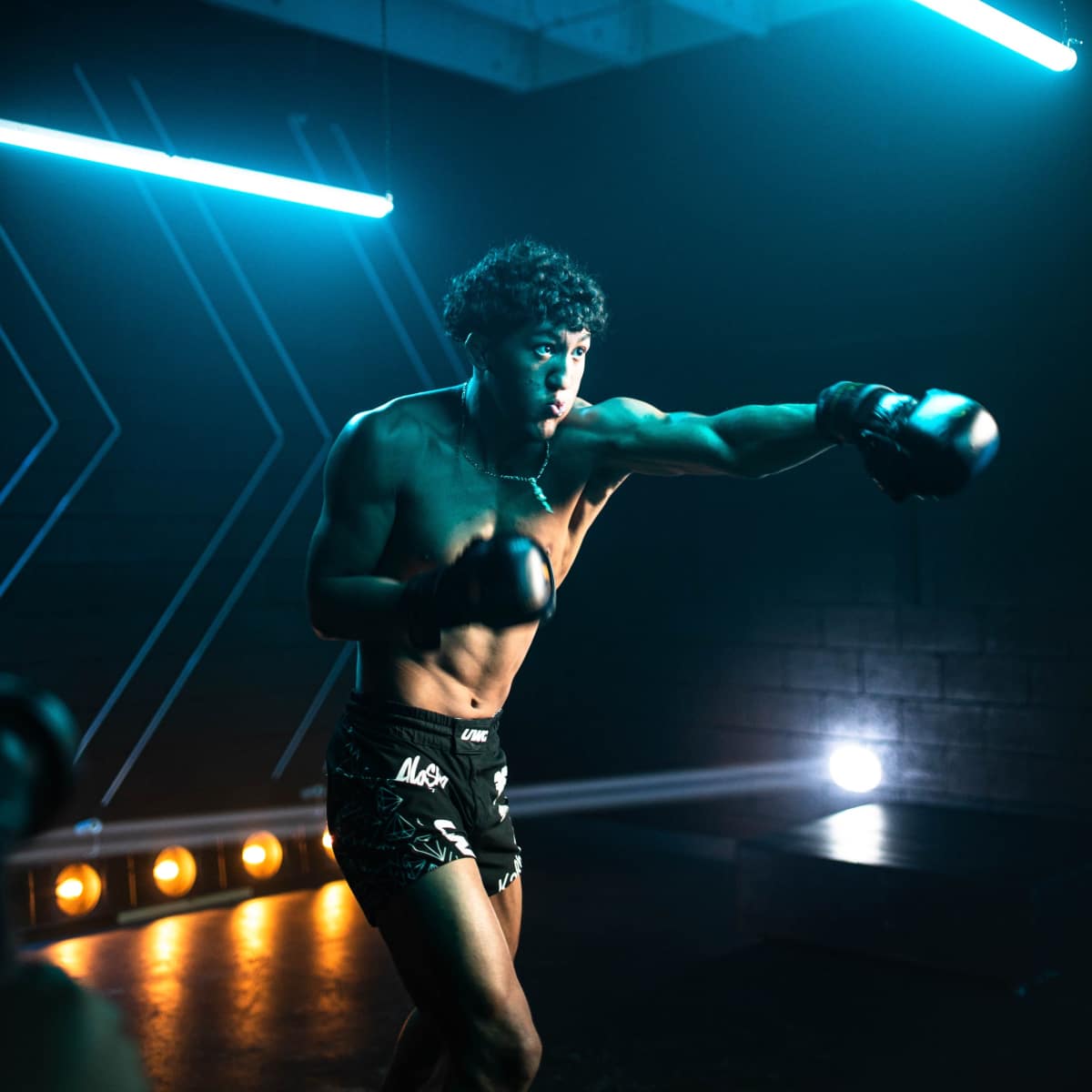 A shirtless boxer throws a punch while wearing gloves in a dimly lit gym with blue neon lighting.