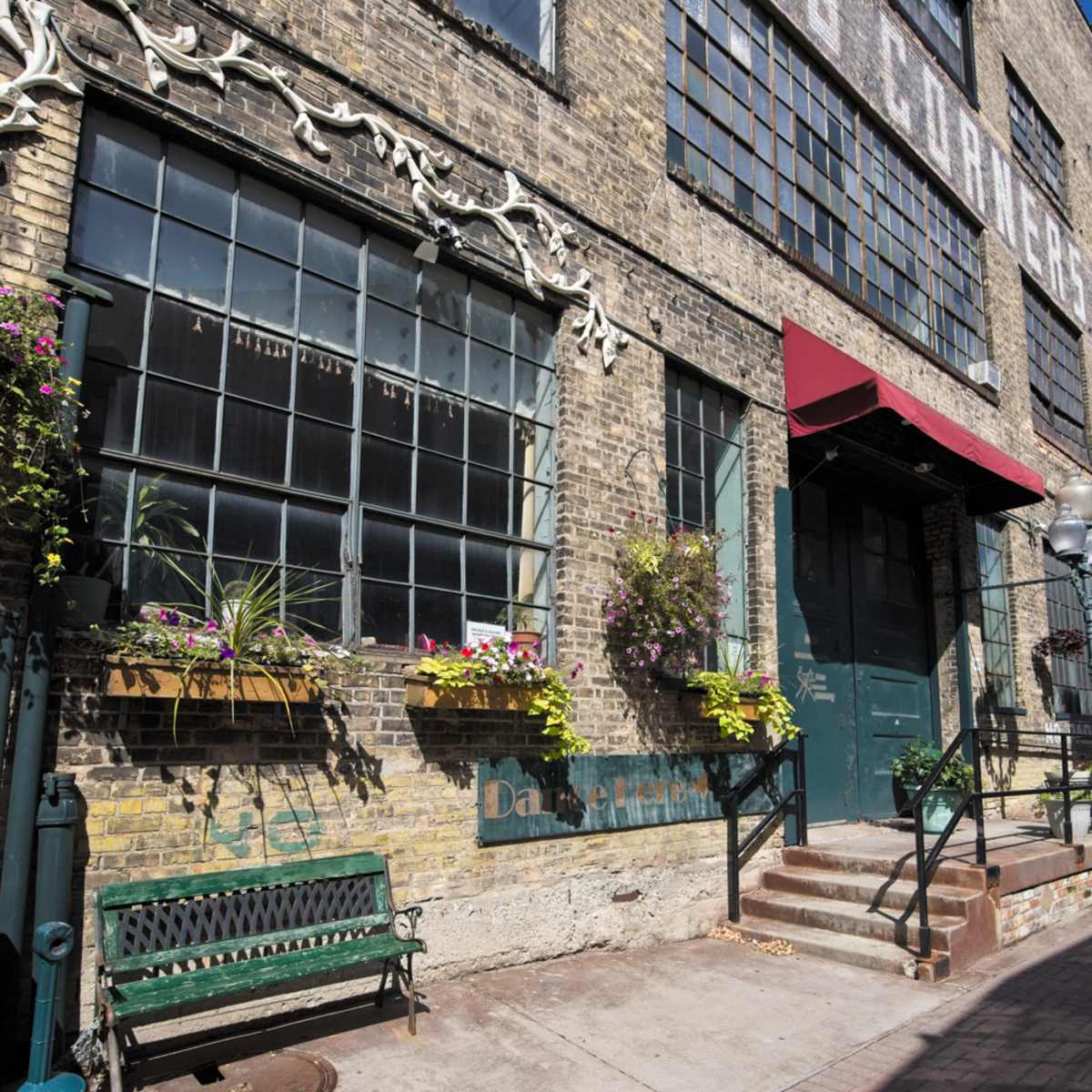 A row of brick buildings featuring large windows and awnings, with planters filled with flowers along the lower level and a concrete walkway in front.