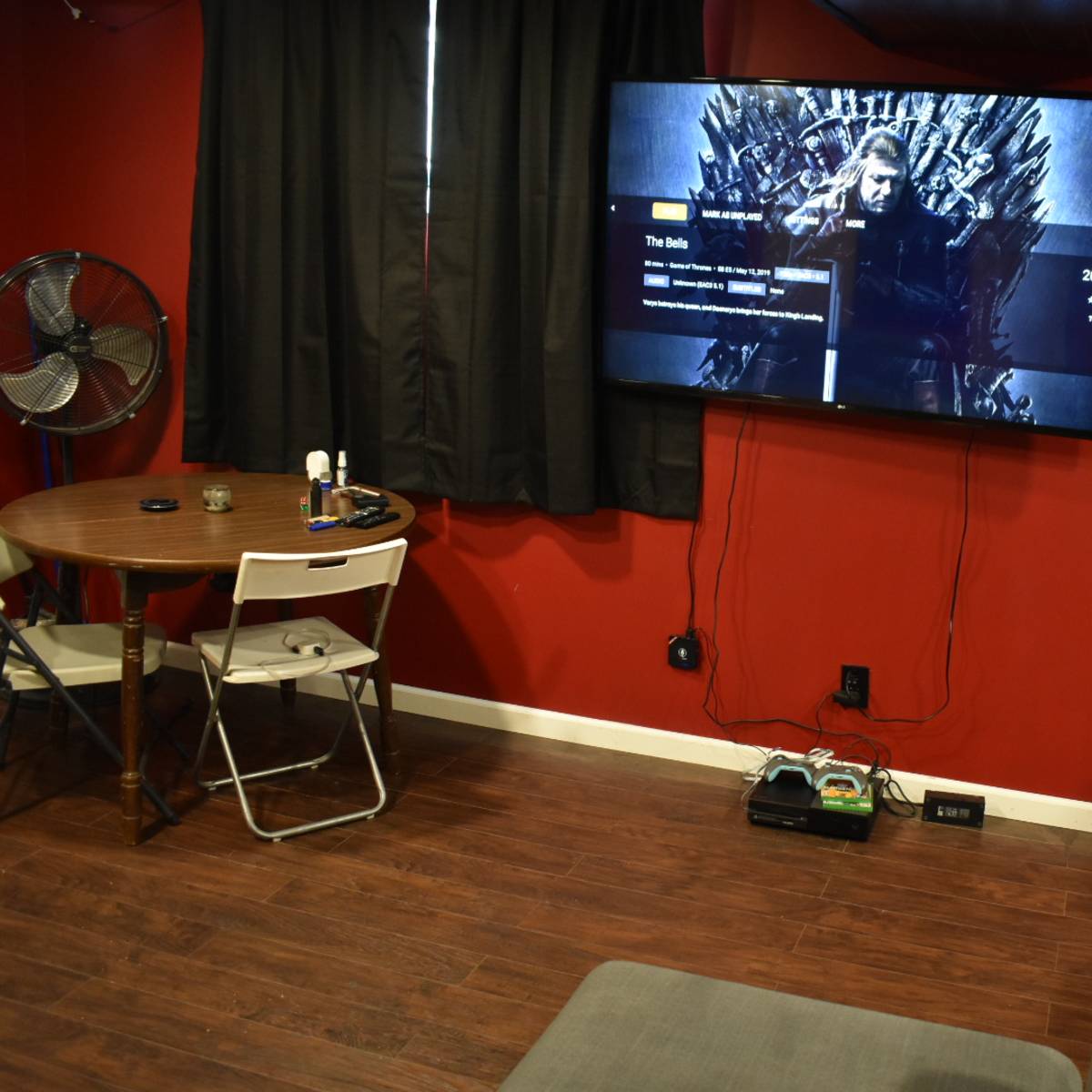 A room with red walls featuring a large television displaying a scene from a show, a round table with chairs, and a fan, with a gray sofa facing the TV.