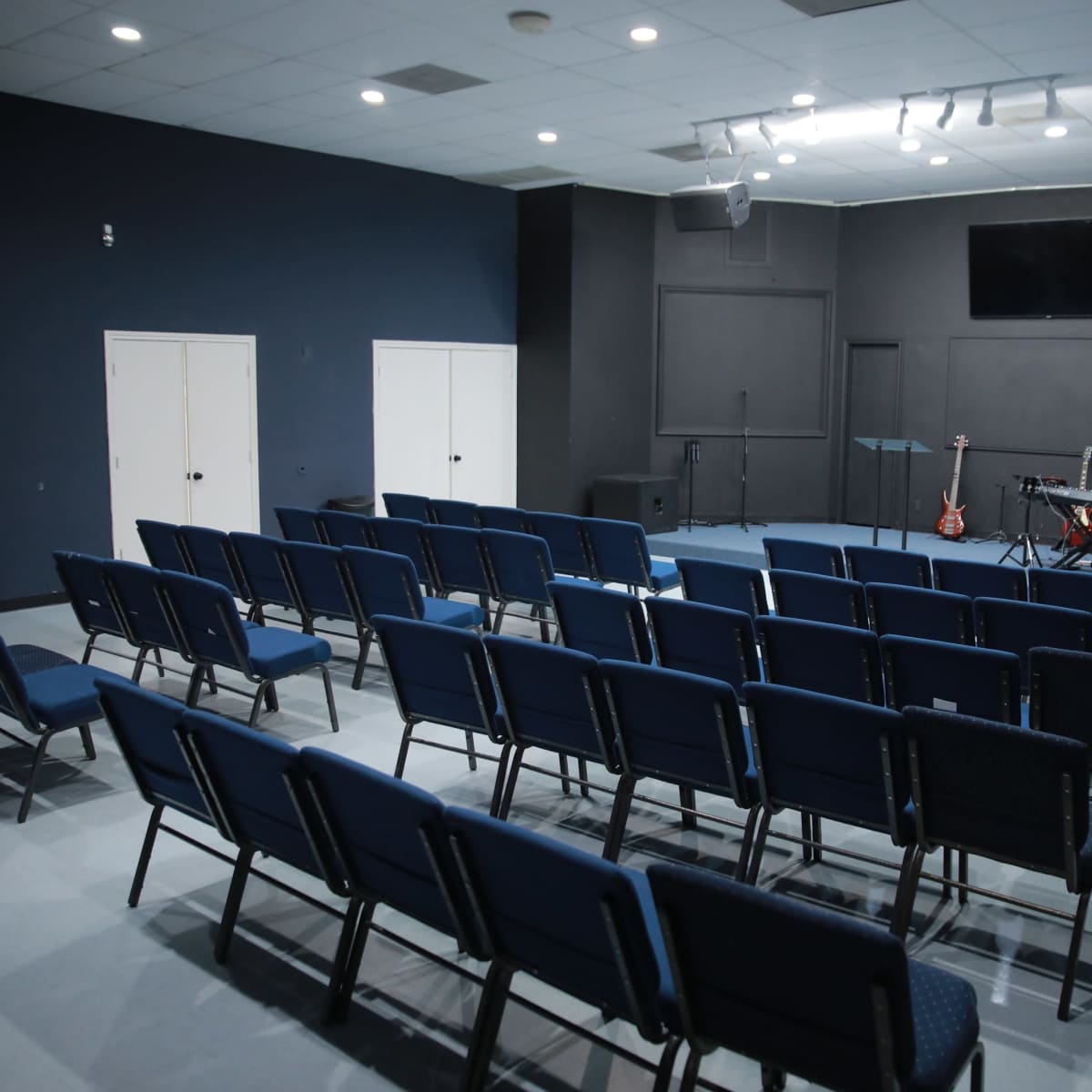 A sparsely arranged indoor space with blue chairs facing a stage equipped with musical instruments.