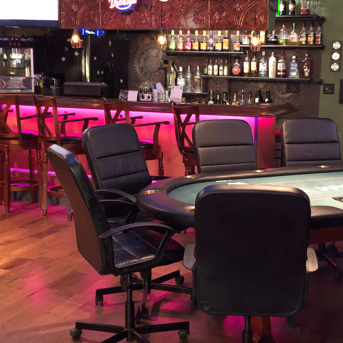 A poker table surrounded by several black chairs in a dimly lit bar setting with a backlit shelf of liquor bottles.