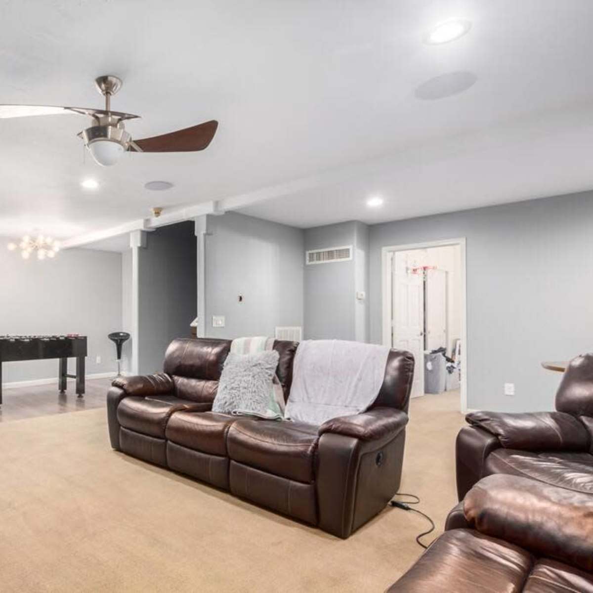 A spacious living room with brown leather sofas, a light-colored carpet, a ceiling fan, and a foosball table.