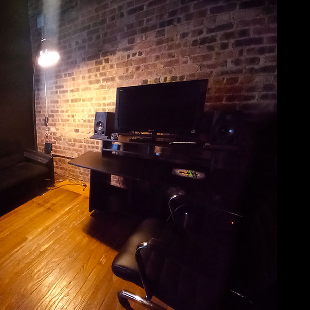 A dimly lit room with a brick wall, a black sofa, a desk with a computer monitor, and audio speakers.