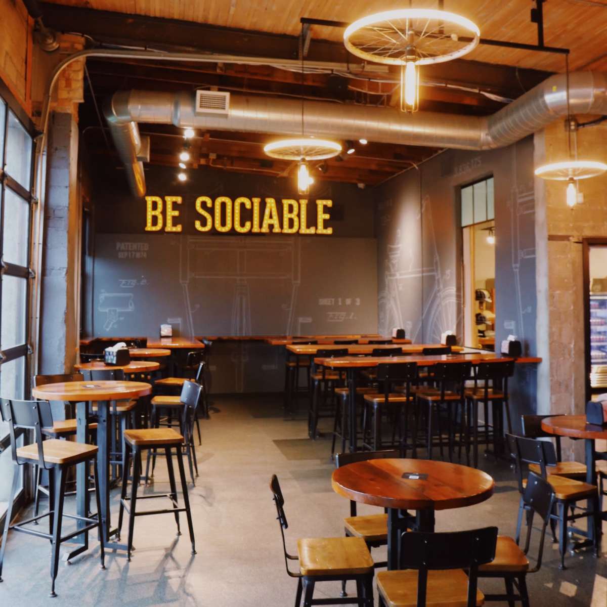 The interior of a café features wooden tables and chairs, with a large sign reading "BE SOCIABLE" on the wall.