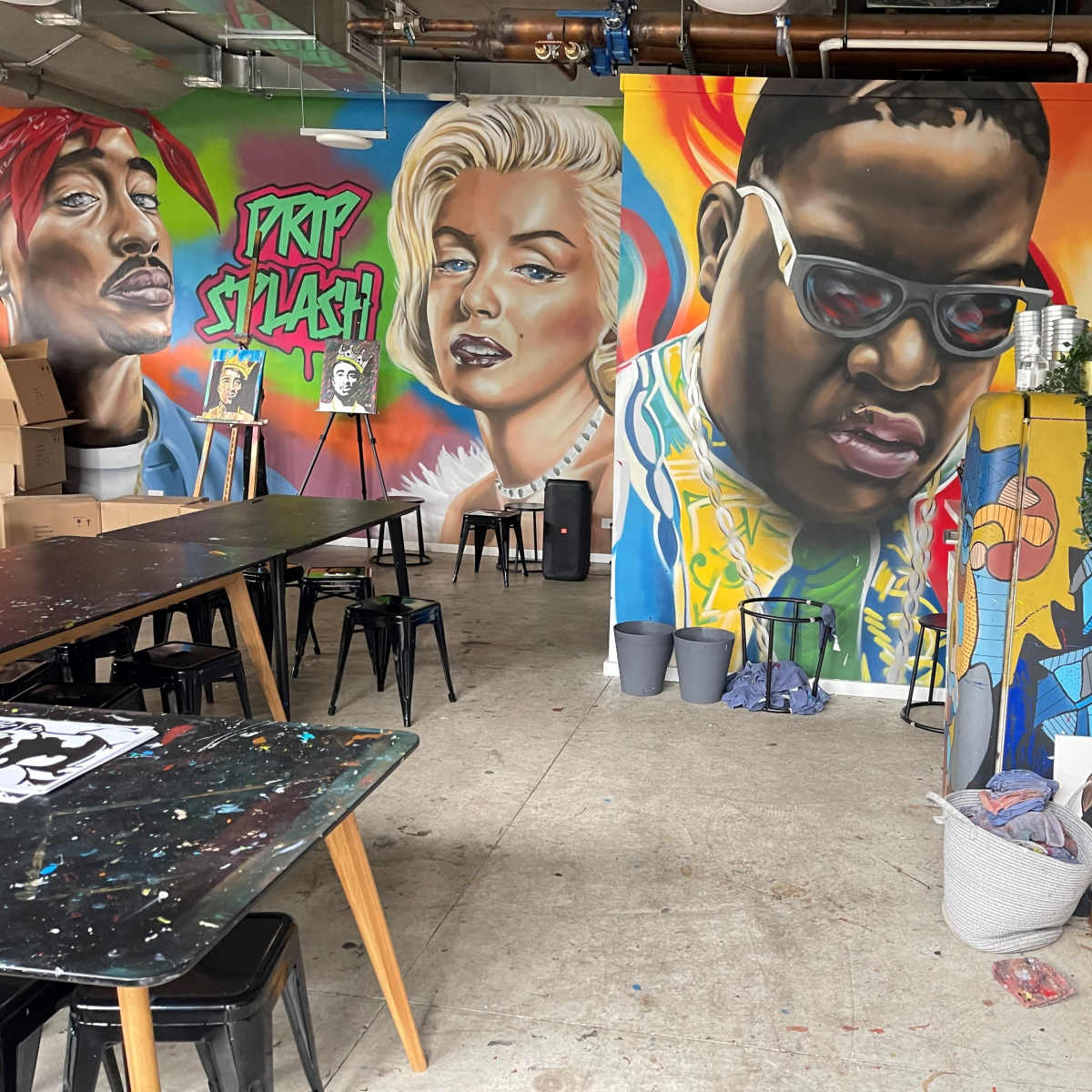 A vibrant art studio featuring large murals of prominent figures on the walls, along with black tables and various art supplies scattered throughout the space.