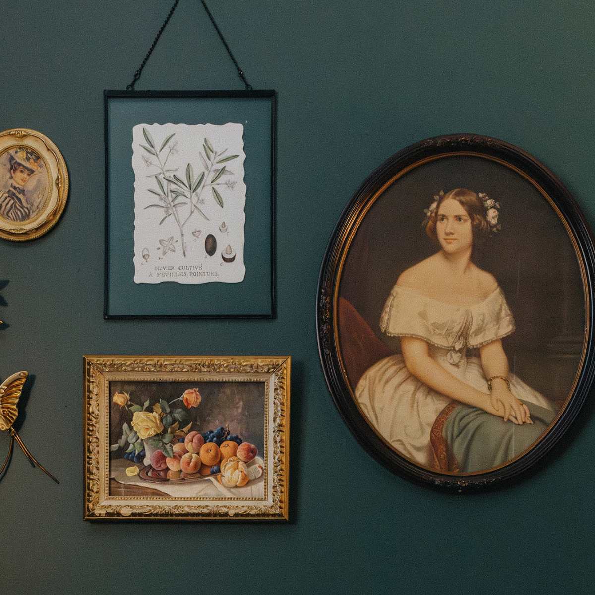 A collection of framed artworks and decorative elements arranged on a green wall, including a portrait of a woman, a botanical illustration, and floral decorations.