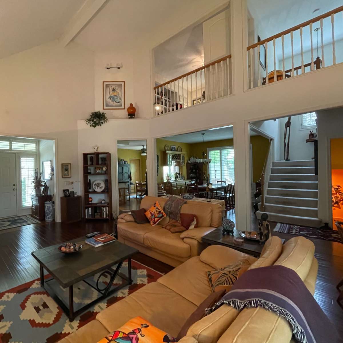 The living room features a beige sofa set, a coffee table, and a large television, creating an open space that connects to a dining area and a staircase.