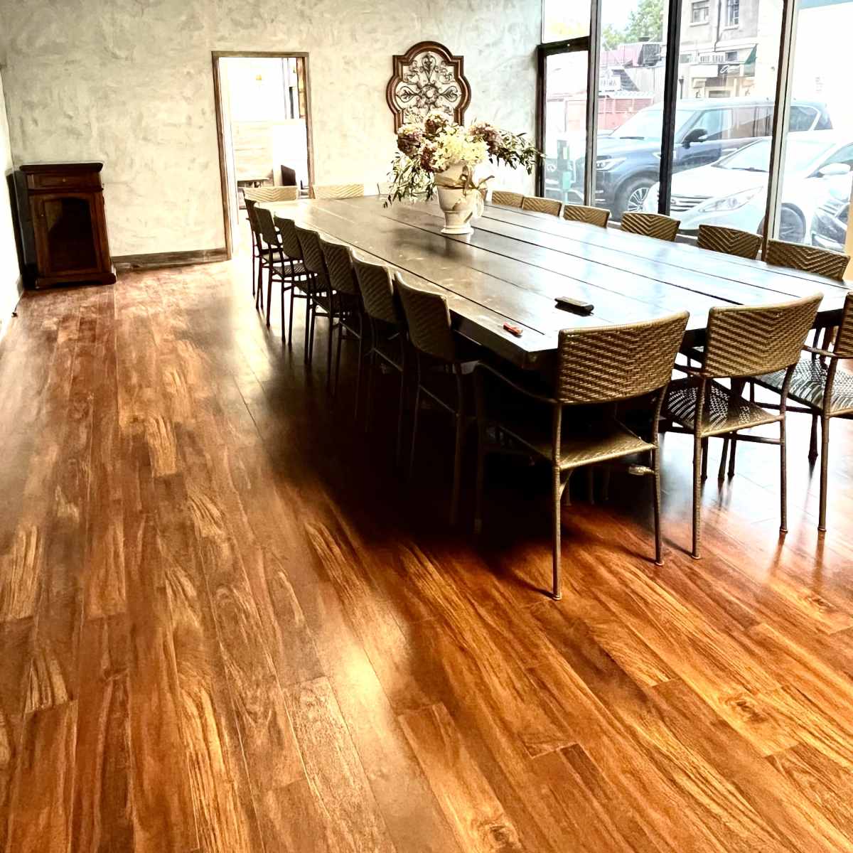 A long wooden table with chairs is set up in a bright room with large windows and a light-colored wall.