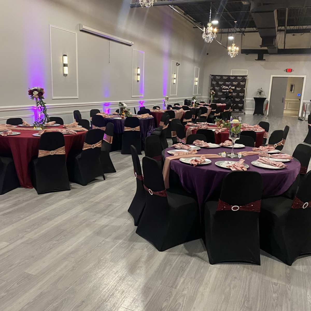 A banquet hall set up with round tables covered in black and burgundy tablecloths, adorned with centerpieces and place settings, under decorative chandeliers.