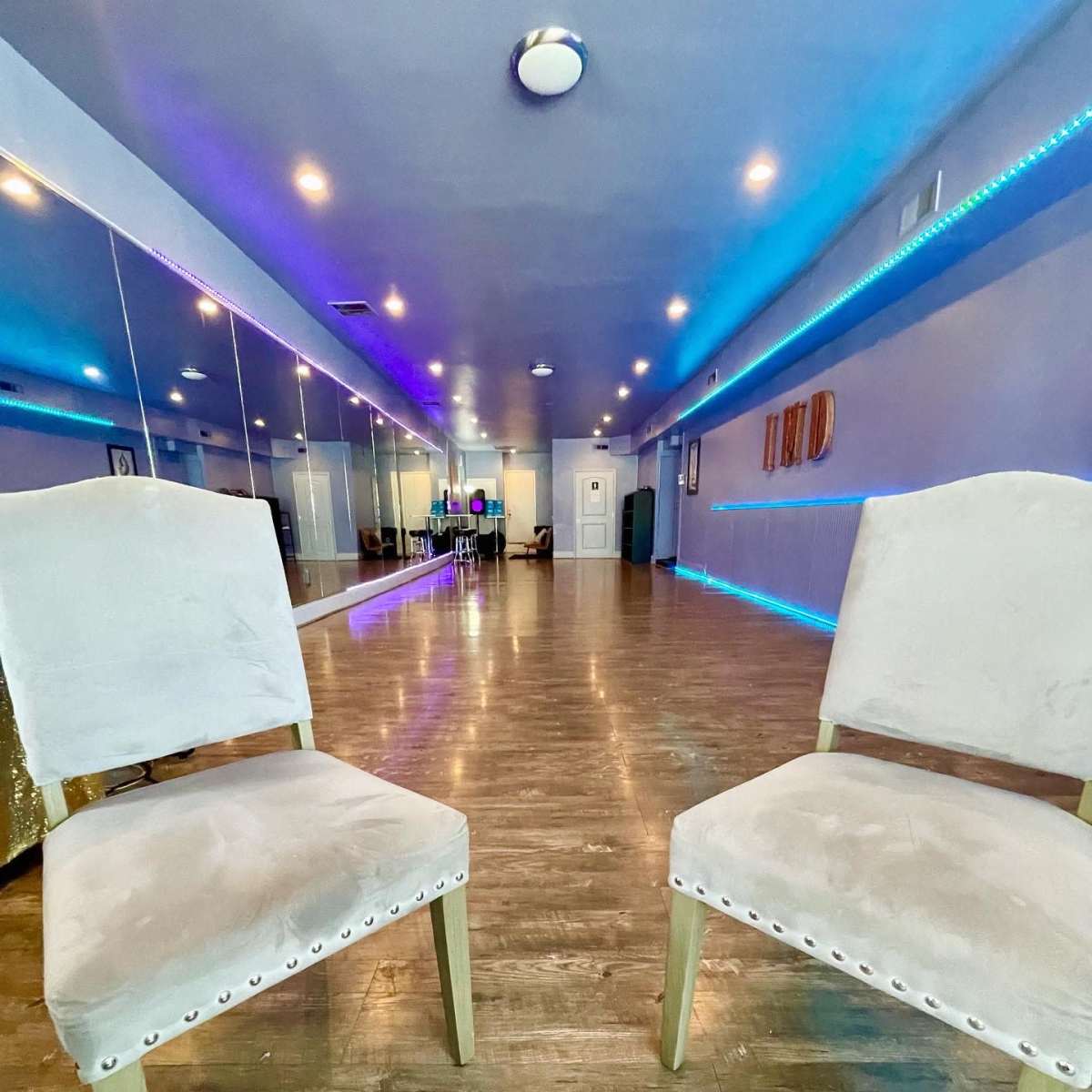 Two upholstered chairs with decorative nails are positioned facing each other in a brightly lit room featuring mirrored walls and colorful LED lights.