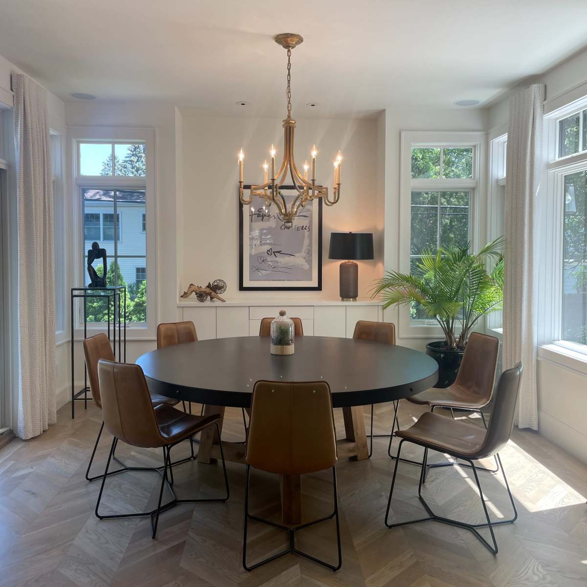 A round dining table with upholstered chairs is illuminated by a chandelier in a well-lit dining room featuring large windows and decorative plants.