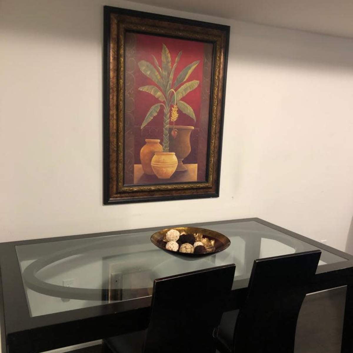 A glass-topped table with two black chairs sits beneath a framed painting of a plant and vases on a white wall.