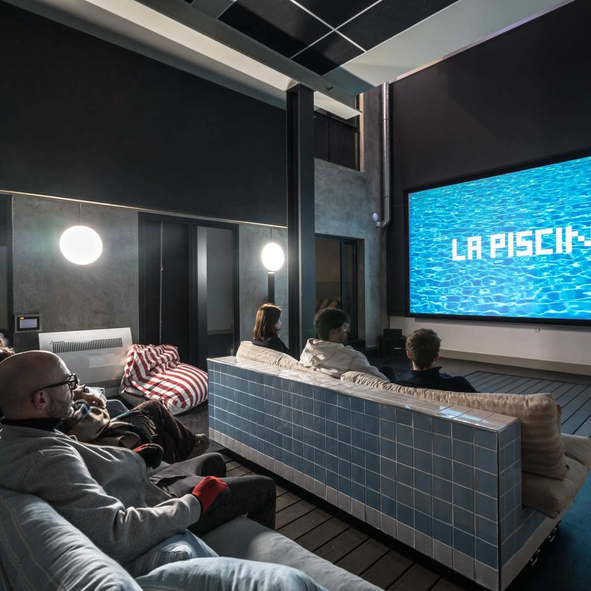 A group of six people sit on sofas in a modern home theater, watching a large screen displaying the words "LA PISCINE" over a blue water background.