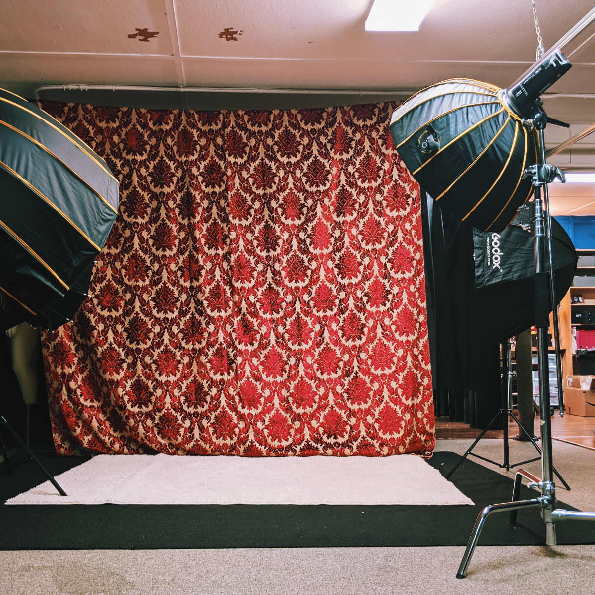 A photography studio setup featuring two large softbox lights positioned in front of a decorative red patterned backdrop and a white rug on the floor.