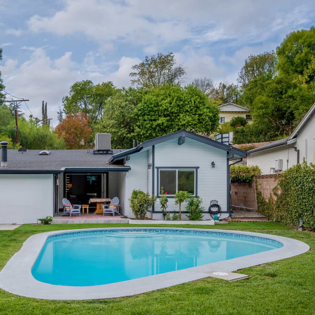 A backyard features a swimming pool surrounded by green grass, lounge chairs, and a patio area with a table and umbrella.