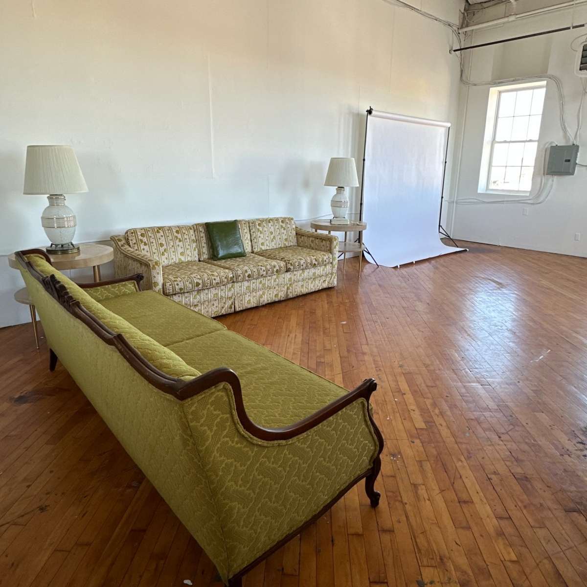 A spacious room with wooden floors featuring a long, ornate green sofa and two matching lamps next to it.