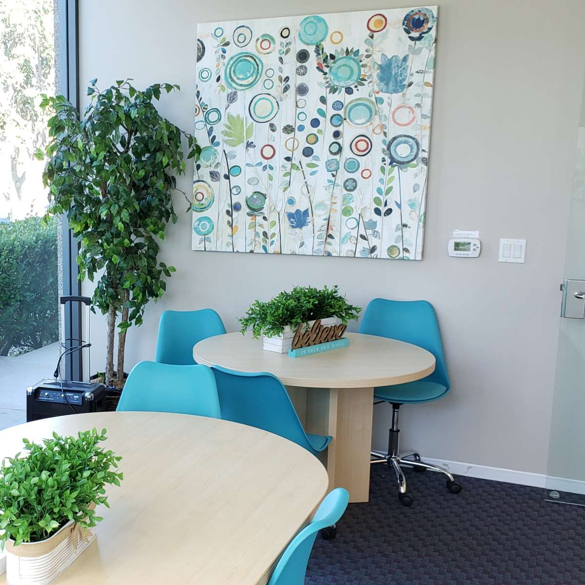 A modern conference room with two round tables and bright blue chairs, featuring a large artwork on the wall and plants in white pots.